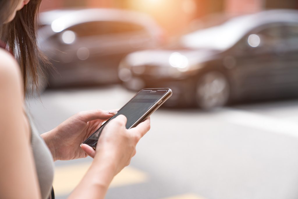 Image of a woman using her smartphone with a parked car in the background for Google Has Launched the First Ride Hailing Price Comparison Tool, That Include Uber and Lyft blog post
